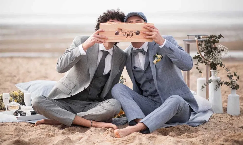 Zwei Männer in Anzügen sitzen barfuß am Strand und küssen sich hinter einem Holzschild mit der Aufschrift Für immer ab jetzt