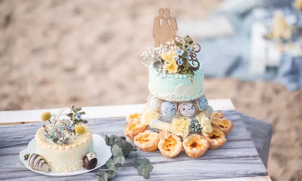 Zwei festlich dekorierte Hochzeitstorten mit Blumen und Tuxedo-Erdbeeren, die größere mit männlichem Paar-Topper, serviert am Strand